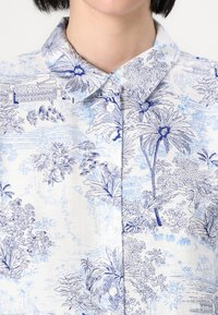 Cămașă ușoară cu un material alb cu modele florale albastre, guler clasic și închidere cu butoane. Suprafață texturată cu desene intricate.