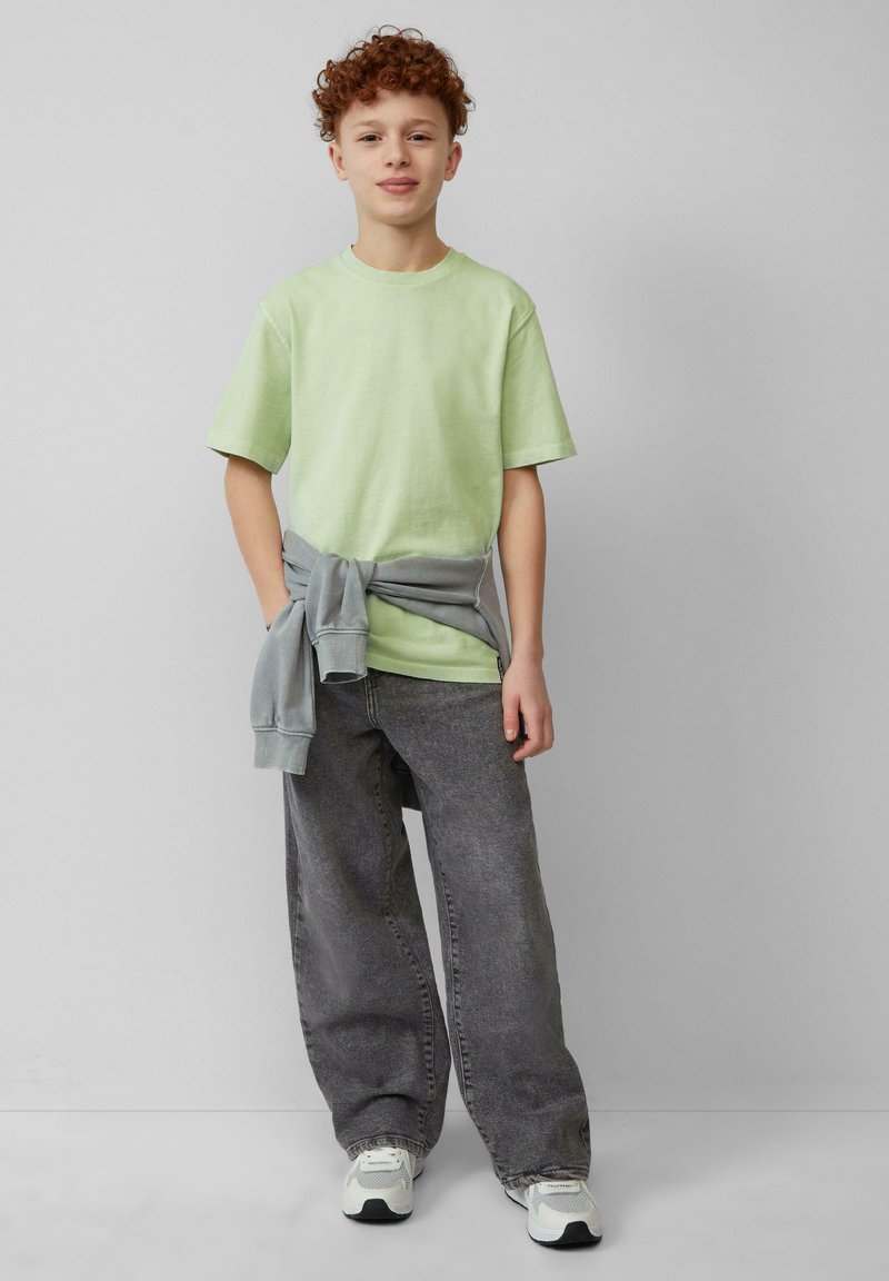 Lichtgroen T-shirt met korte mouwen, grijze sweatshirt geknoopt om de taille, losse grijze jeans en witte sneakers. Gladde stof, ontspannen pasvorm.