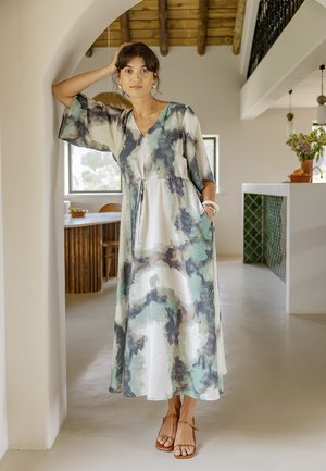 Donna in un abito midi fluido con motivo astratto e maniche a gomito, in piedi in una stanza moderna e luminosa con soffitto in legno e arco.