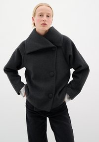 Dunkelgraue Wolljacke mit breitem Kragen, großen Knöpfen und lockerer Passform. Graue Bündchen sind unter den Ärmeln sichtbar.