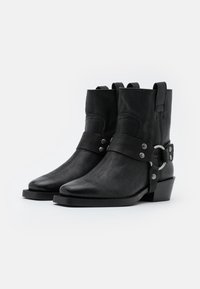 Botas de tobillo de cuero negro con un acabado texturizado, punta afilada, tacón de altura media, acento de hebilla de metal y detalles de correa.
