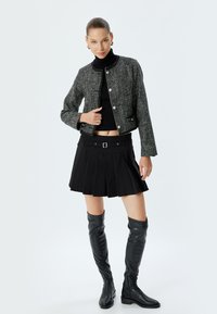 Giacca cropped in tweed nero con bottoni argentati, maglione nero a collo alto, mini gonna plissettata nera e stivali alti in pelle nera fino al ginocchio.