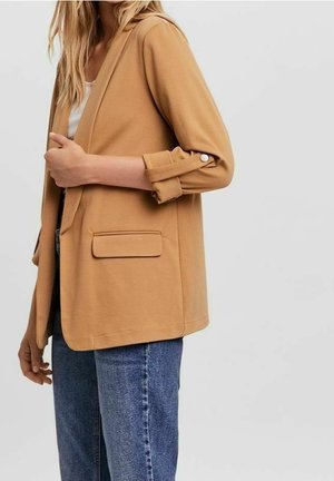 Femme portant un blazer beige aux manches retroussées avec des boutons pression, associé à un jean bleu et un haut blanc, sur un fond uni.