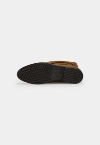 Bottine en suede marron avec un bout rond et une semelle en caoutchouc noir texturé, dotée d'un design de grip à motifs.