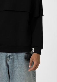 Sweat à col rond noir, superposé, avec une texture. Porté avec un jean en denim bleu clair. Un petit étui noir est tenu dans la main gauche.