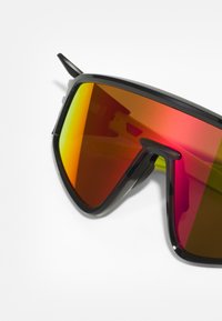 Os óculos de sol apresentam uma grande armação preta angular com lentes refletivas de vermelho-alaranjado e pontas afiadas. O design é desportivo, com uma estética arrojada.
