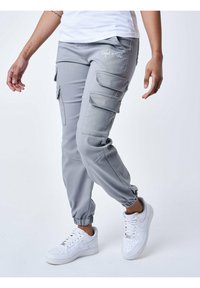 Grijze cargobroek met meerdere zijzakken en elastische enkelmanchetten, gemaakt van een gladde, lichte stof. Gepaarde met witte sneakers.