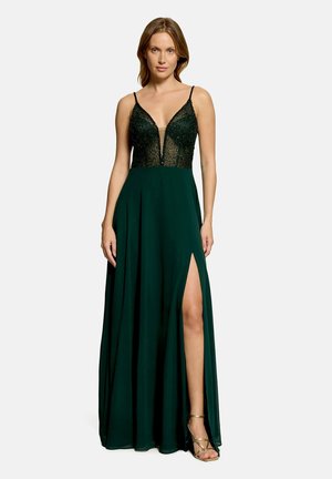Femme portant une robe de soirée vert foncé sans manches, avec un profond décolleté en V, un corsage perlé, une fente haute à la jambe et des sandales dorées à talons hauts à brides.