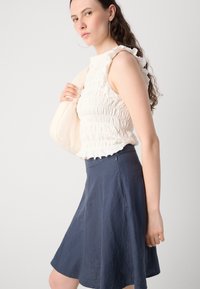 White, sleeveless, smocked top with ruffled shoulders paired with a dark blue flared skirt. The model holds a light-colored bag.