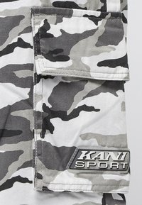 Tkanina s kamuflážním vzorem v odstínech šedé a bílé s klopovou kapsou. Obsahuje logo KANI SPORT ve stříbrném kovovém provedení.