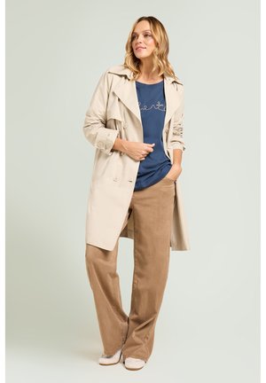Donna in piedi con trench beige, camicia blu scuro, pantaloni marrone chiaro a gamba larga e scarpe bianche su uno sfondo chiaro e uniforme.