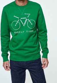 Zelená mikina s jednoduchou bílou grafikou bicyklu a textem "WHEELY TYRED", kterou nosí osoba s tmavými džíny.