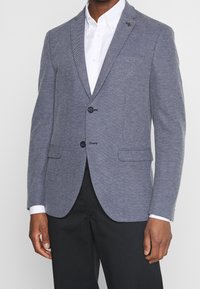 Man som bär en texturerad grå blazer över en vit skjorta med knappar och svarta byxor, stående mot en enkel bakgrund.
