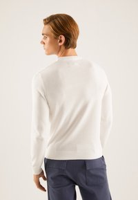 Pull en tricot blanc à col rond et manches longues, présentant une texture lisse et des poignets en côtes, porté avec un pantalon bleu marine.