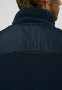 Mörk marinblå fleecejacka med en mjuk textur, som har en slät nylonöverkant på kragen. Designen inkluderar sydda sömmar och en ren, minimalistisk stil.