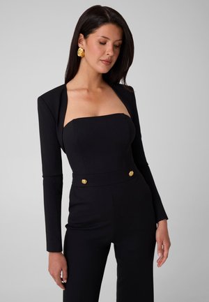 Mujer con cabello largo y oscuro que lleva un mono negro sin tirantes con mangas largas y botones dorados y pendientes, mirando ligeramente hacia abajo.