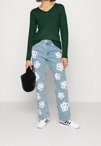 Grüner V-Ausschnitt-Pullover, hellblaue Jeans mit weißen Blumenapplikationen, schwarzer Hut, weiße Sneaker mit schwarzen Streifen. Lässiges Outfit-Design.