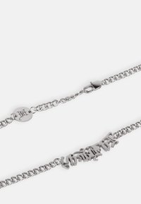 A pulseira de corrente de prata apresenta elos curvos interligados com uma placa decorativa que exibe texto estilizado e um fecho de lagosta.
