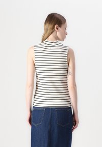 Bele sleeveless, prigludęs viršus pagamintas iš lengvo audinio, su tamsiai mėlynais ir baltais horizontaliais raštais, aukštu kaklu ir subtilia logotipo detale nugaros pusėje.