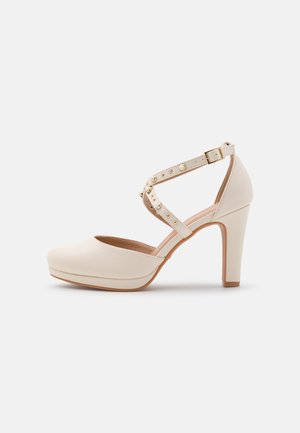 Cream-colored high-heeled shoe with a rounded toe, adjustable ankle strap, and gold stud accents on the strap. Smooth texture.