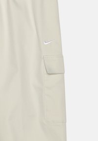 Svetlo béžové cargo šortky s výraznou bočnou kapsou a malým vyšívaným strieborným logom Nike na hornej strane. Hladká textúra.