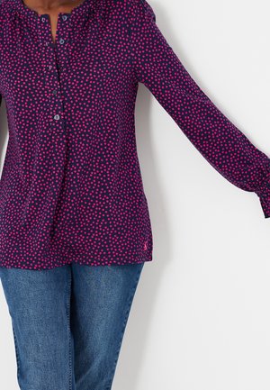 Kvinde iført en navyblå bluse med lange ærmer og lyserøde polkaprikker samt blå jeans, stående med den ene arm strakt ud mod en hvid baggrund.