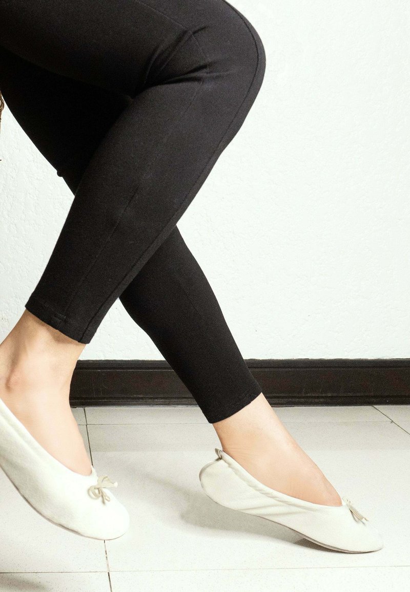 Ballerines en tissu blanc avec un bout arrondi, détail de nœud noué, associées à un legging noir ajusté, présentées sur un fond de sol clair.