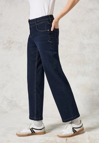 Donkerblauwe, wijde jeans met een hoge taille, voorzakken, zichtbare stiksels en een aansluitende tailleband, gecombineerd met witte sneakers en grijze sokken.