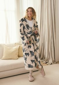 Nő mosolyog bézs színű köntösben, amelyen fekete bika minták vannak, miközben megköt egy övet, egy bézs kanapé mellett áll párnákkal teli, világos nappaliban.