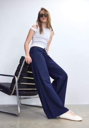 Junge Frau trägt ein weißes Cold-Shoulder-Top, marineblaue Hosen mit weitem Bein, weiße Ballerinas und Sonnenbrille, sitzt auf einem modernen schwarzen Ledersessel.