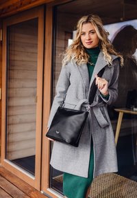 Černá kožená kabelka přes rameno s hladkým povrchem a stříbrnými doplňky, kterou drží osoba v šedém kabátu a zelených šatech.