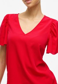 Sarkana V-veida blūze ar pūkām īsām piedurknēm, izgatavota no viegla auduma, gludas tekstūras un minimālisma dizaina bez rotājumiem.