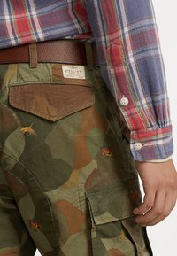 Pantalones cargo de camuflaje con diseños de insectos bordados en colores vibrantes. El material es de algodón, con un bolsillo de parche marrón y costuras resistentes.