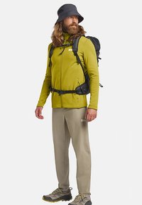 Giacca in pile verde, pantaloni beige, zaino nero e scarpe da trekking grigie. Caratterizzata da un design strutturato e un cappello a secchiello.