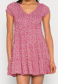 Mujer vestida con un vestido rojo de manga corta, con un patrón floral de margaritas blancas y un escote en V, de pie contra un fondo liso.