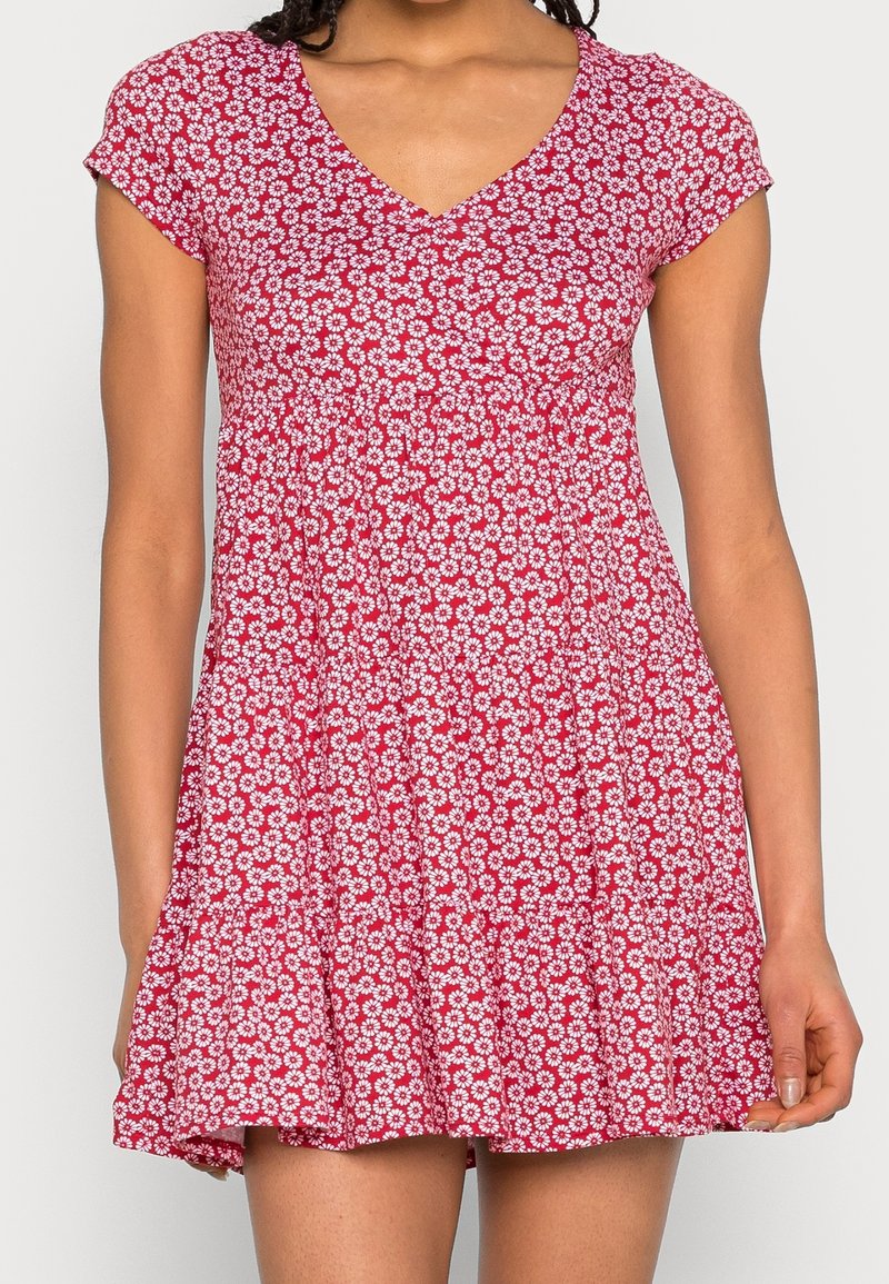 Mujer vestida con un vestido rojo de manga corta, con un patrón floral de margaritas blancas y un escote en V, de pie contra un fondo liso.