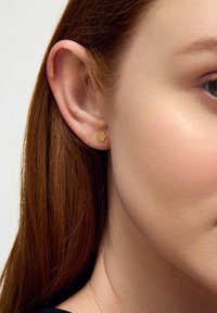 Złoty sześciokątny kolczyk wkręcany wyeksponowany na małżowinie usznej, uzupełniający gładką, lekko opaloną cerę oraz proste, rudawe włosy.
