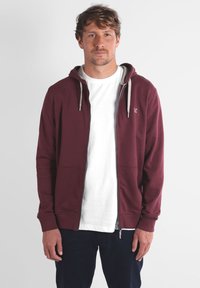Sweat à zip bordeaux avec un cordon gris, deux poches avant et un t-shirt blanc à col rond en dessous ; tissu doux, design décontracté.