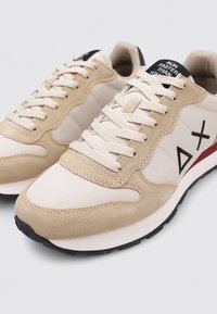 Baskets de course beige et crème avec des lacets épais, des accents en daim, une semelle noire et un logo noir sur le côté, posées sur un fond blanc.