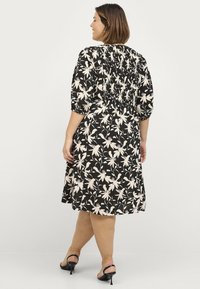 Schwarzes Kleid mit einem weißen Blumenmuster, elastischer Smocking hinten, kurzen Ärmeln und einem ausgestellten Rock, getragen mit schwarzen Absätzen.