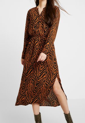 Mujer que lleva un vestido de manga larga con estampado de cebra en naranja y negro con una abertura lateral, combinado con botines de color verde oscuro.