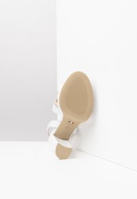 Sandale à talons blancs avec des lanières larges, tige en cuir lisse et semelle en caoutchouc beige présentant un motif texturé. Étiquette "Fabriqué en Italie" visible.