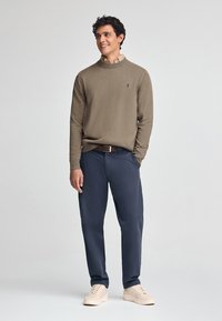 Maglione a maglia marrone con colletto, pantaloni blu navy e sneakers crema. Presenta un logo discreto sul maglione e una cintura marrone.