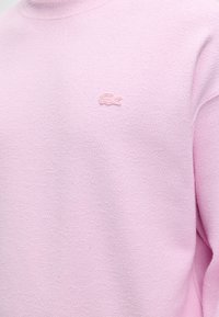 Ανοιχτό ροζ υφασμάτινο φούτερ με στρογγυλή λαιμόκοψη και μικρό κεντητό ροζ λογότυπο κροκοδείλου στην αριστερή πλευρά του στήθους.