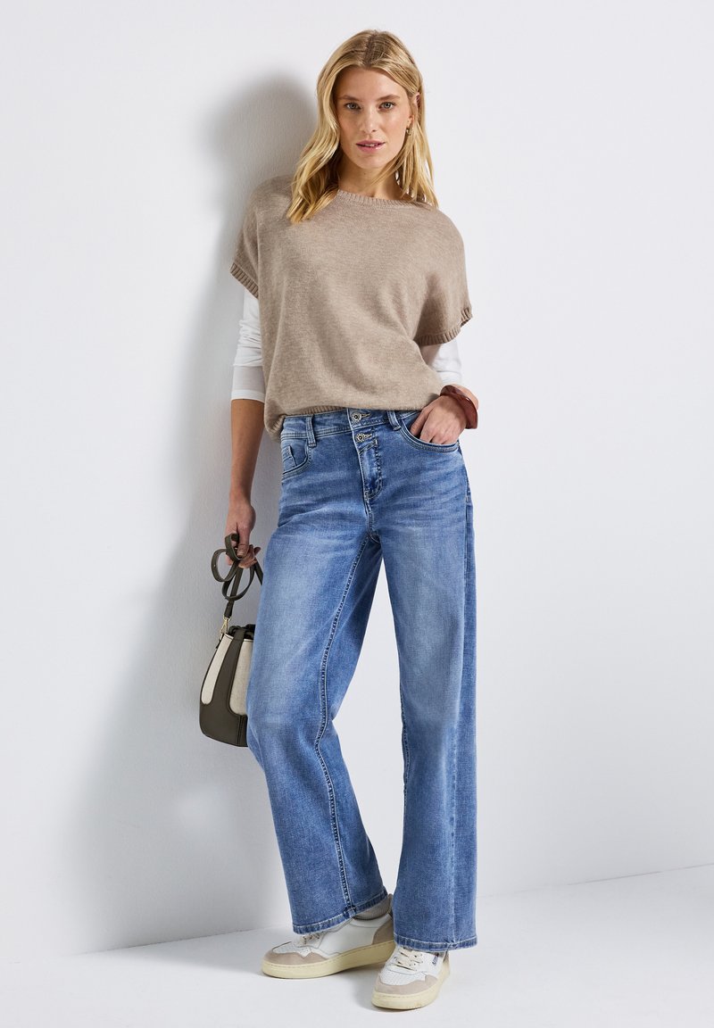 Beige gebreide top met korte mouwen, gecombineerd met lichtblauwe wijde jeans. Geaccentueerd met een kleine handtas en witte sneakers.