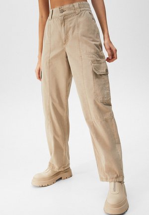 Beige cargo broek met wijde pijpen en voor- en zijzakken, gedragen met bijpassende stevige ritslaarzen, getoond op een model van taille tot voeten.