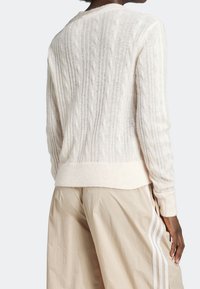 Suéter de punto trenzado crema con dobladillo y puños de canalé. Presenta un patrón texturizado y un sutil brillo. Se lleva con pantalones beige claros.