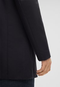 Navy blazer met een getailleerde pasvorm, gladde textuur, met notched revers en een enkele knoopsluiting. De mouwen hebben strakke lijnen en geen versieringen.