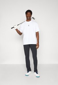 Hvid bomulds t-shirt med et lille grafisk logo, parret med mørkegrå bukser og hvide golf sko med blå detaljer. Holder en golfkølle.