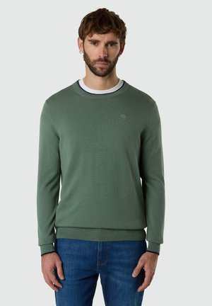 Mann mit Bart, der einen grünen Pullover über einem weißen Hemd und blauen Jeans trägt und vor einem einfarbigen hellen Hintergrund steht.